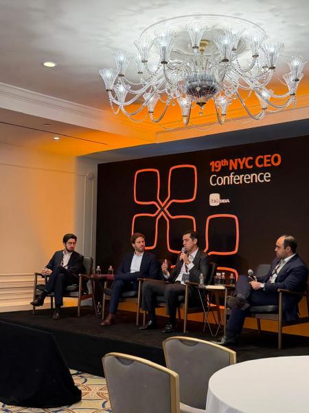 Painel de discussão no 19º NYC CEO Conference, com quatro homens falando em um cenário moderno e elegante, sob um lustre.