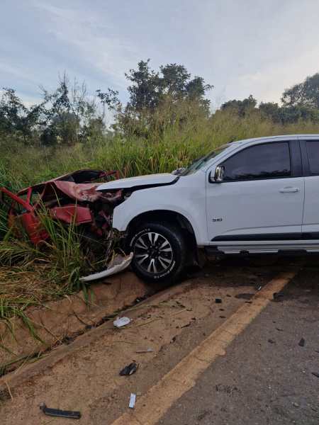 Veículo branco colidido em um acidente, próximo a um carro vermelho danificado em um terreno gramado.
