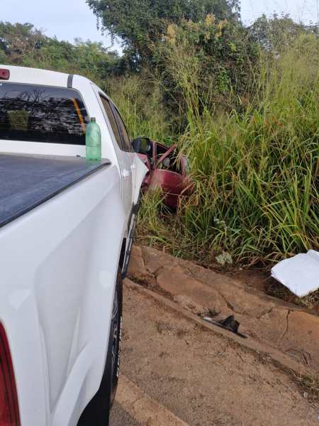 Um carro vermelho parcialmente escondido em vegetação alta, visto ao lado de uma caminhonete branca.