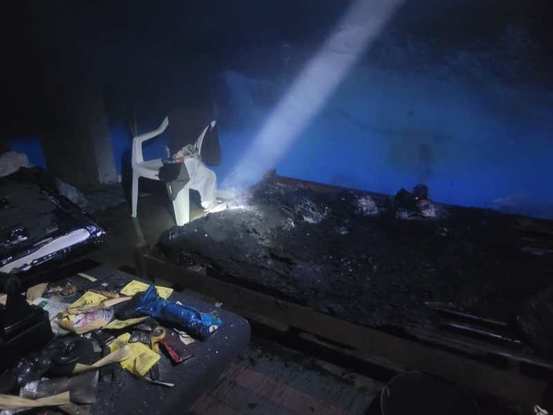 Interior de um ambiente escuro, com sinais de incêndio. Uma cadeira branca está próxima de objetos queimados e uma luz formando um feixe. O chão está coberto de detritos.
