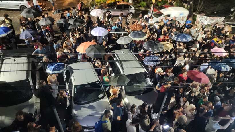 Multidão de pessoas com guarda-chuvas se reunindo em uma área pública, cercada por carros, em uma noite chuvosa.