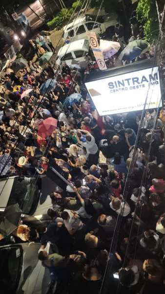 Multidão reunida na rua, com pessoas segurando guarda-chuvas, próximo a um outdoor do SINTRAM, à noite.