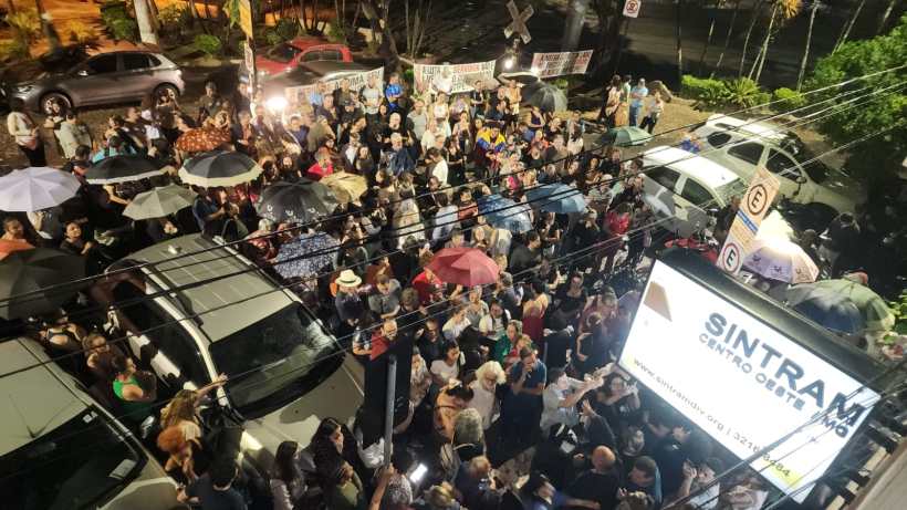 Multidão de pessoas em uma noite, algumas segurando guarda-chuvas, em uma rua com carros estacionados e placas visíveis.