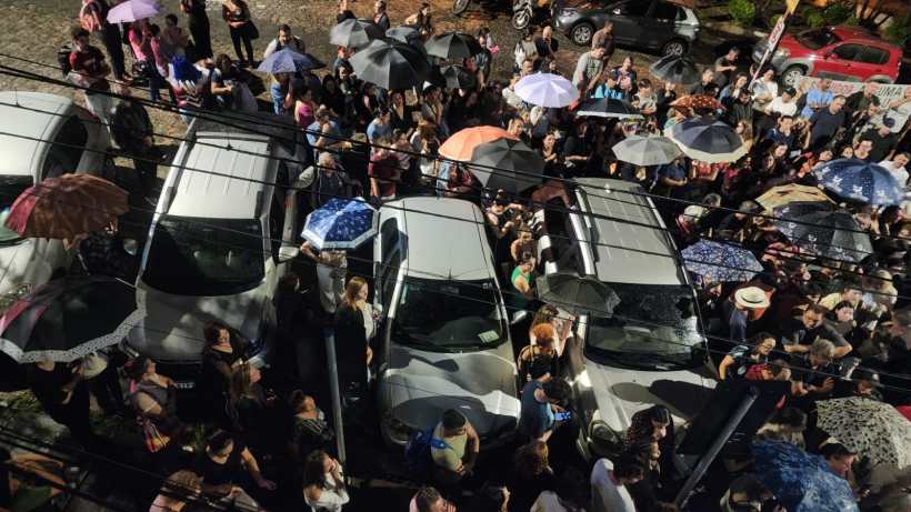 Multidão de pessoas com guarda-chuvas em uma rua, cercando carros estacionados.