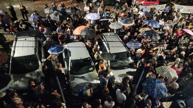 Multidão de pessoas com guarda-chuvas em uma rua, rodeando carros estacionados.