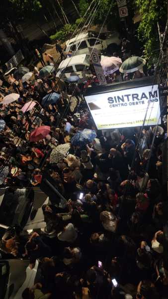 Multidão de pessoas reunidas sob guarda-chuvas em uma rua, com um letreiro iluminado do SINTRAM em destaque.