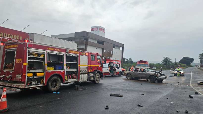 Acidente de trânsito com veículos danificados na estrada, com caminhão dos bombeiros e veículos de emergência presentes ao lado de um hotel.