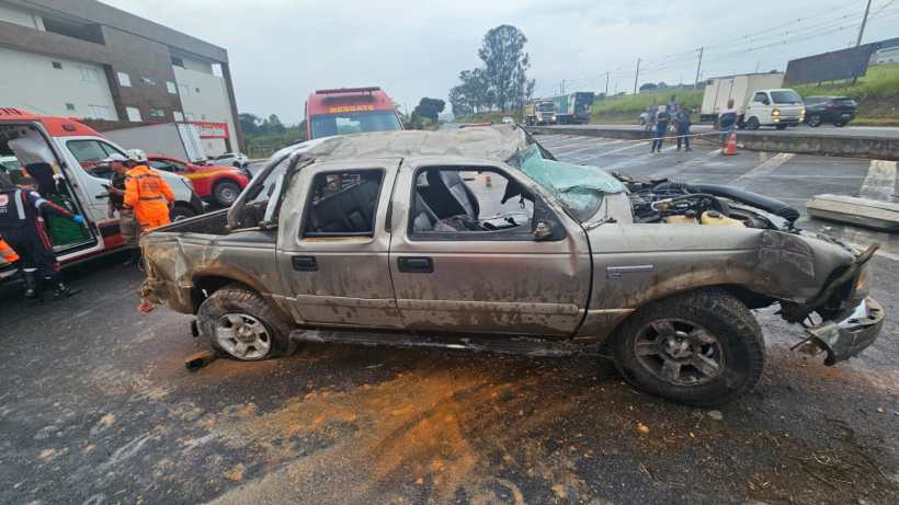 Veículo danificado em um acidente, com uma lateral amassada, na estrada, ao lado de ambulâncias e equipes de resgate.