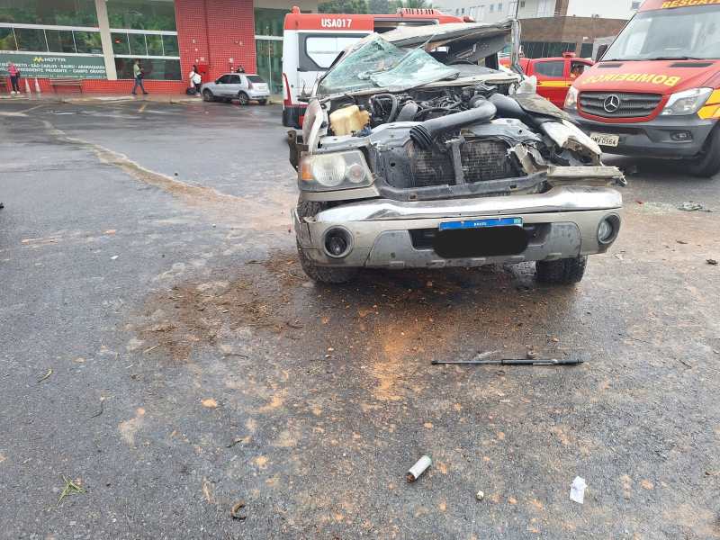 Carro acidentado com capô danificado e motor exposto em área urbana, com marcas de óleo no chão.