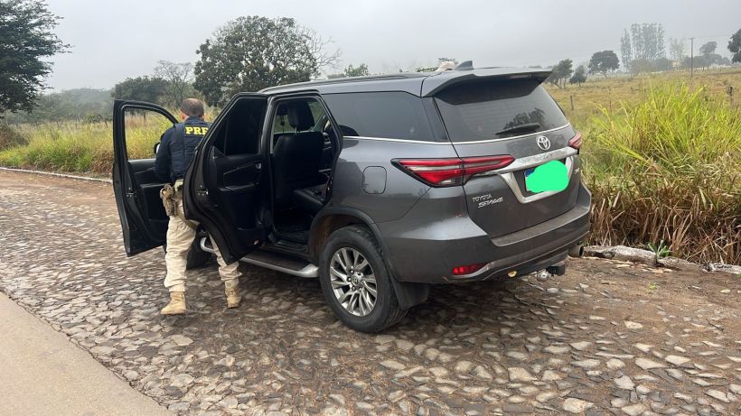 Veículo SUV Toyota estacionado em uma estrada de paralelepípedos, com uma pessoa vestindo uniforme da PRF (Polícia Rodoviária Federal) perto do carro, que está com as portas abertas.