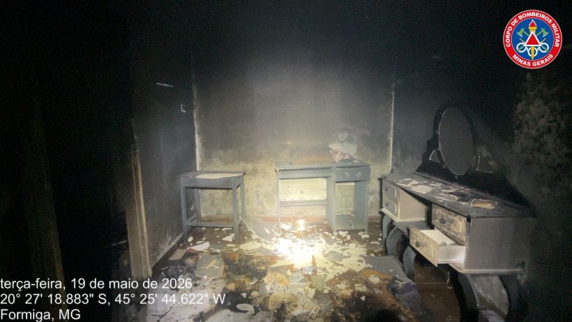 Interior de um ambiente danificado por incêndio, com móveis carbonizados e resíduos no chão. Luz fraca iluminando a cena.