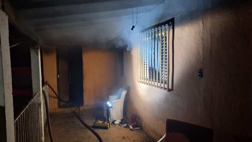 Área externa de uma casa com fumaça, iluminação fraca e móveis visíveis, incluindo uma cadeira e um tubo de mangueira.