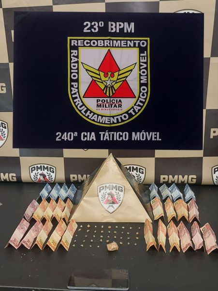 Imagem de uma mesa com material apreendido pela polícia, incluindo uma pirâmide feita de notas de dinheiro, com uma bandeira do 23º BPM ao fundo e várias notas organizadas ao redor.