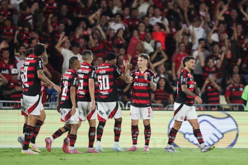 A group of football players celebrating a goal, wearing red and black striped jerseys. The background shows enthusiastic fans in a stadium.