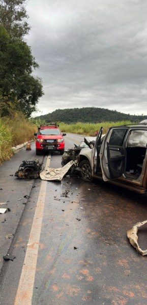 Acidente de carro em uma estrada com um veículo destruído e outro carro vermelho ao fundo.