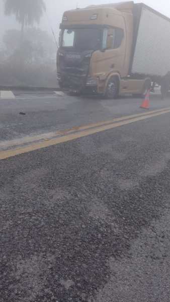 Caminhão parado em uma estrada com neblina, parcialmente coberto por uma lona, e um cone de sinalização ao lado.