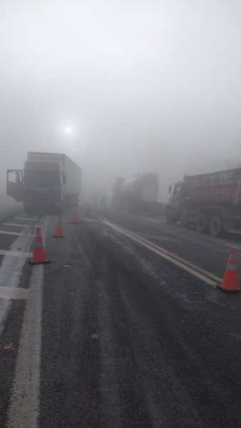 Caminhões parados em uma estrada nevoenta, com cones de sinalização laranja visíveis.