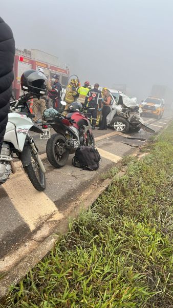 Acidente de trânsito com veículos danificados, incluindo motos e um carro. Equipe de resgate e bombeiros trabalhando na cena em um dia com névoa.