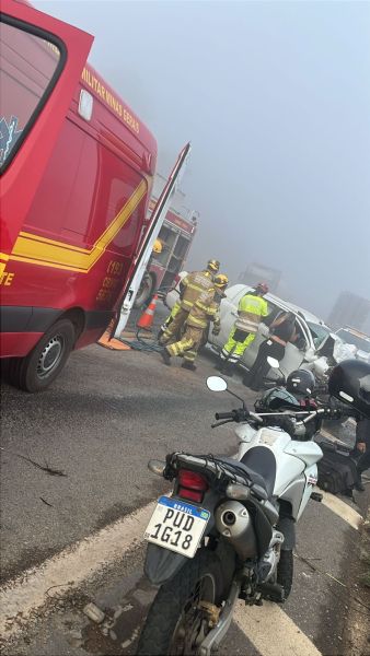Acidente de trânsito em uma rodovia com visibilidade reduzida devido à neblina. Equipes de resgate e bombeiros atendem vítimas em um veículo danificado, enquanto uma motocicleta está estacionada ao lado.