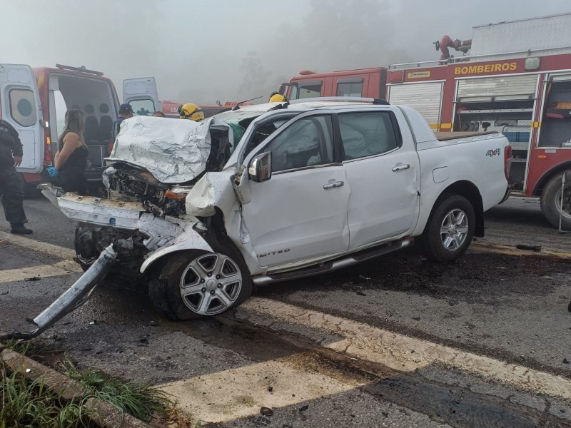 Acidente de carro com uma picape branca danificada, mostrando a parte da frente esmagada. Há ambulâncias e bombeiros ao fundo em uma estrada com neblina.