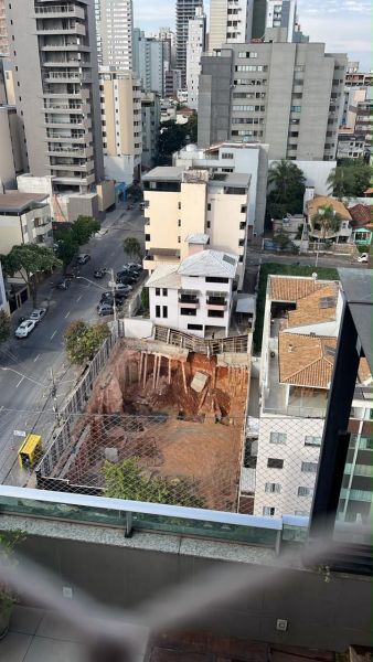 Vista aérea de uma área de construção em uma cidade, com um grande buraco escavado em terra, cercado por estrutura de metal, cercado por prédios e ruas.