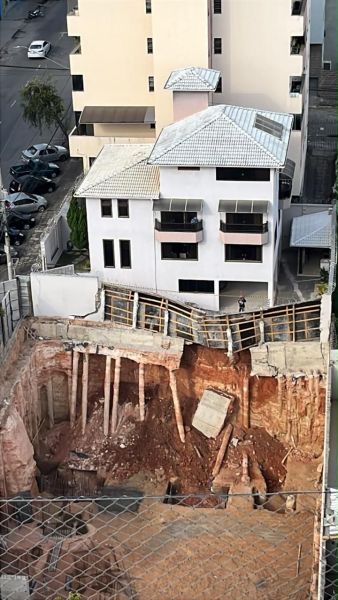 Imagem de uma obra de construção com uma escavação profunda, revelando estruturas de madeira de suporte, em frente a um edifício residencial.