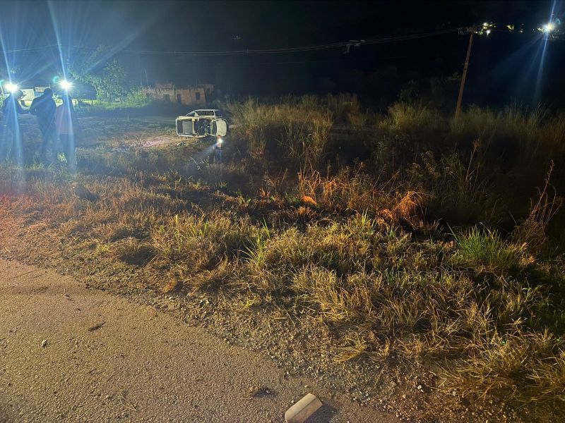 Veículo capotado em um campo durante a noite, com luzes de emergência e pessoas ao redor.
