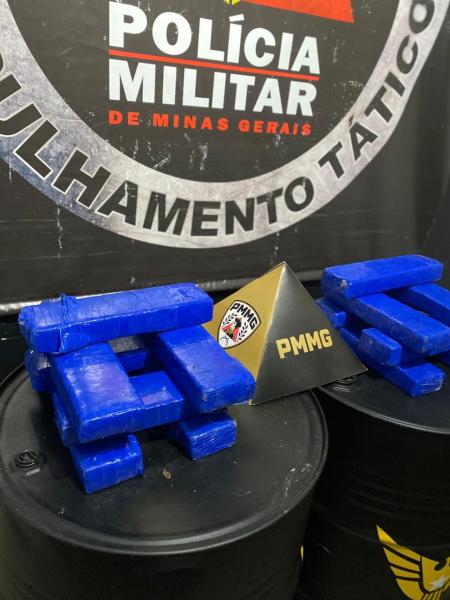 Bloquinhos de cor azul dispostos em uma mesa, com um triângulo dourado com a sigla PMMG ao centro, em frente a barris pretos, contra um fundo de parede com o emblema da Polícia Militar de Minas Gerais.