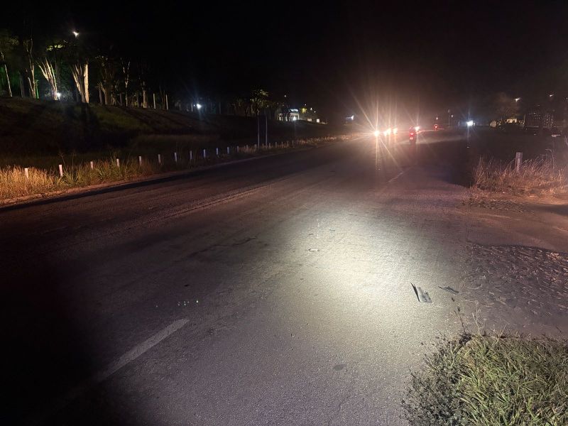 Uma estrada iluminada à noite, com vegetação nas laterais e veículos se aproximando ao fundo.