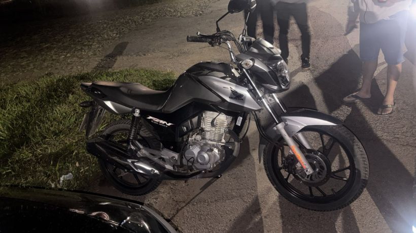 Moto Honda CG 160 Fan prata em um espaço externo à noite, com pessoas ao fundo.
