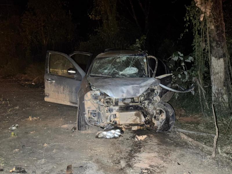 Carro acidentado em uma área escura, com danos significativos na frente e janelas quebradas. A porta do carro está aberta, e há detritos no chão ao redor.