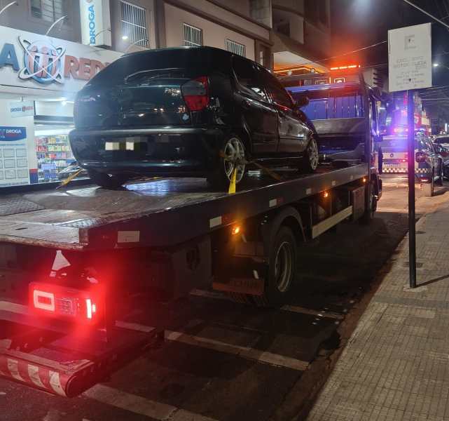 Um carro preto sendo rebocado por um caminhão guincho à noite, em uma rua urbanizada.