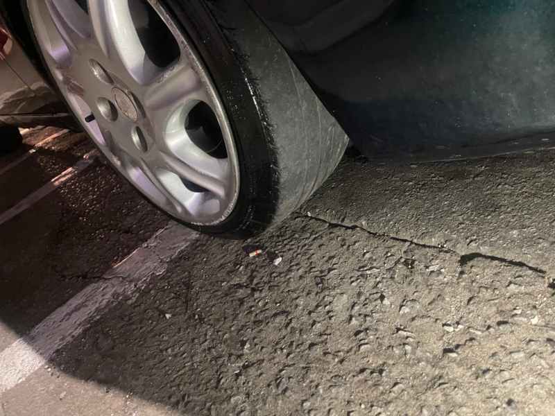 Pneu de carro furado em estacionamento durante a noite, com foco na roda e na superfície da estrada.