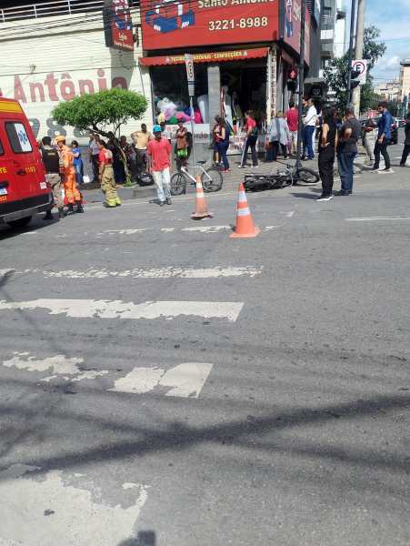 Cena de um acidente na rua com um motociclista caído e pessoas reunidas ao redor. Uma ambulância e bombeiros estão presentes, enquanto cones de sinalização estão posicionados na faixa de pedestres.