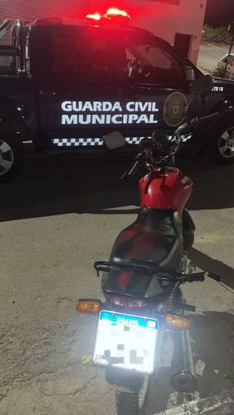 Motocicleta vermelha com placa visível em primeiro plano, e viatura da Guarda Civil Municipal ao fundo com luzes de emergência acesas.