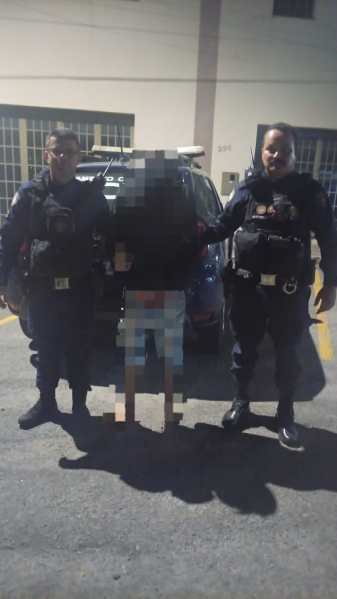 Dois policiais em pé ao lado de um homem com as mãos para trás, em frente a um carro da polícia.
