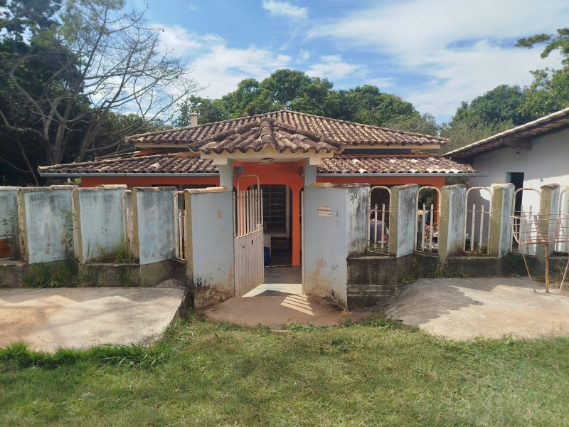 Casa de estilo rústico com telhado de cerâmica, cercada por um muro baixo. A entrada tem um portão aberto, e ao fundo há vegetação.