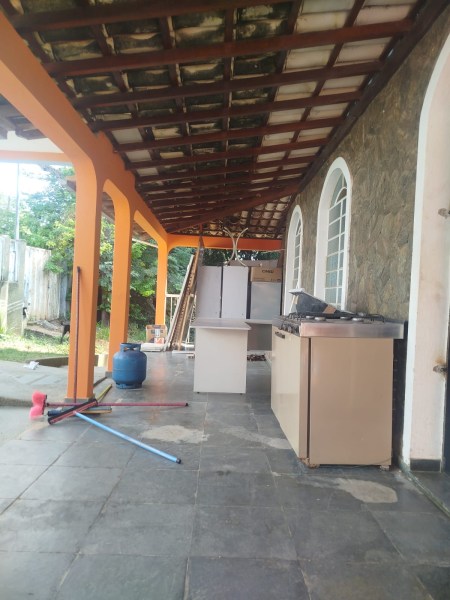 Varanda de uma casa com piso de pedra, apresentando uma cozinha externa com fogão e pia, além de um tanque e utensílios de limpeza ao fundo. A estrutura tem colunas laranjas e um telhado de madeira.