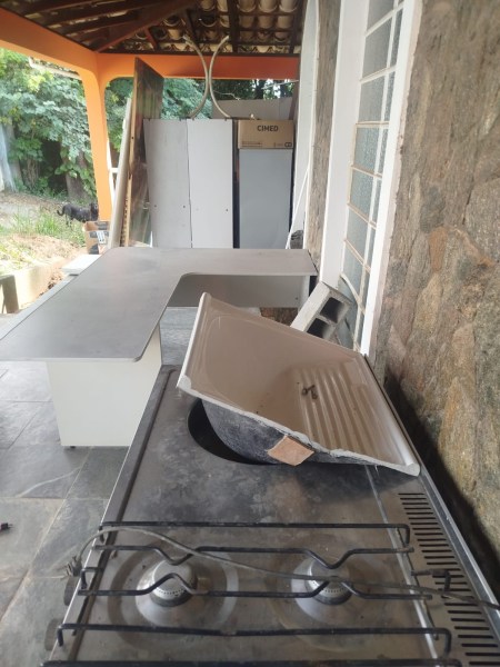 Cozinha externa com uma pia e um fogão, revestida de pedra, com uma bancada em L ao fundo e armários brancos.