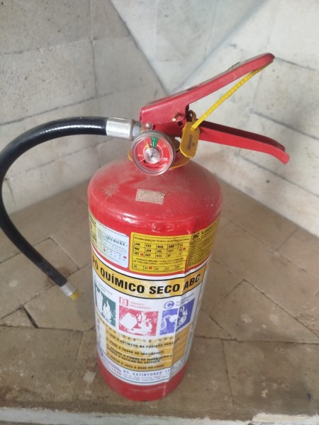 Extintor de incêndio tipo químico seco, com bico e manômetro visíveis, posicionado em uma superfície de alvenaria.