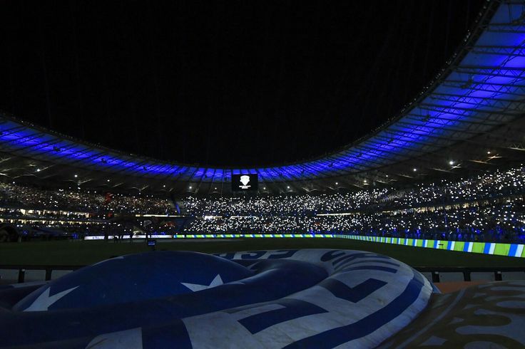A stadium at night filled with blue lighting, showcasing fans holding their phones in the stands, while a large flag is visible in the foreground.