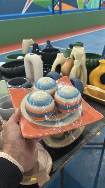 Um homem segurando um prato laranja com três tigelas coloridas, em um ambiente com várias cerâmicas e utensílios de cozinha ao fundo.