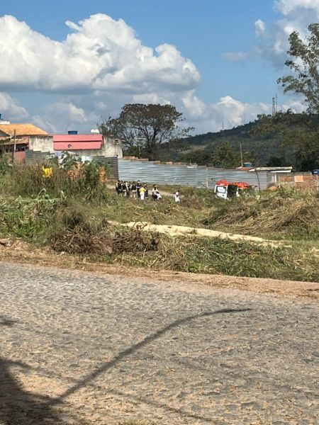 Grupo de pessoas em uma área aberta, com vegetação e barracos ao fundo, céu azul com nuvens.