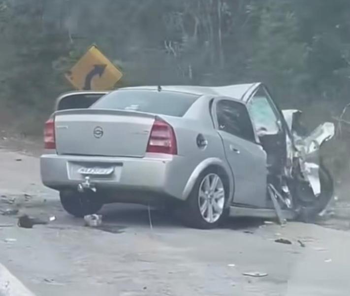 Um carro prata acidentado na beira da estrada, com danos visíveis na porta do lado direito e uma placa de sinalização de curva ao fundo.