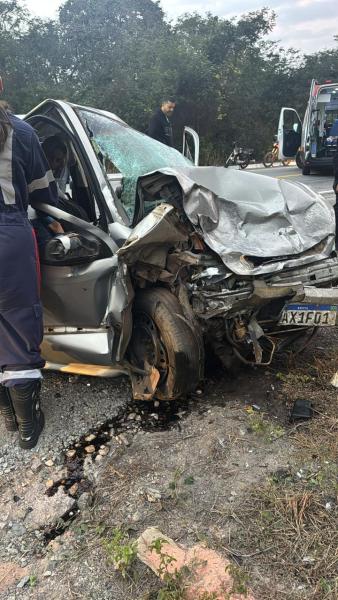 Carro danificado em acidente com a parte frontal esmagada, resíduos de óleo na estrada e pessoas auxiliando na cena.