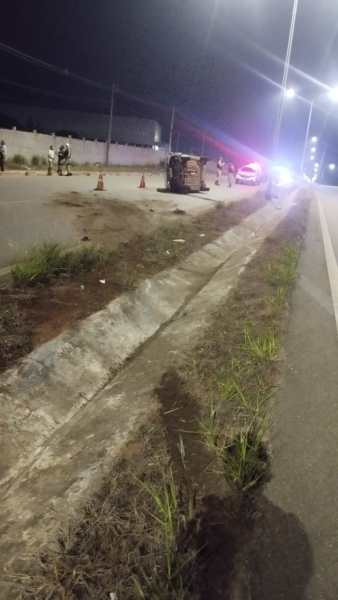 Caminho à noite com um carro capotado ao lado da estrada, com sinalização de emergência e presença de pessoas.