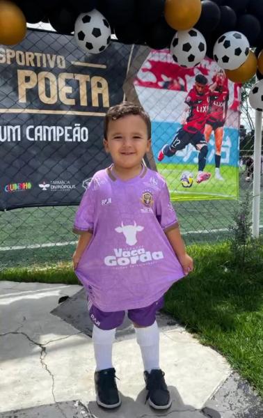 A young child wearing a large pink soccer jersey, standing on a field with a soccer-themed backdrop featuring players and balloons.