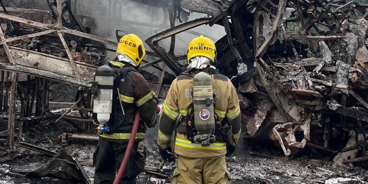 Dois bombeiros em frente a destroços de um incêndio, observando a cena com fumaça ainda visível.