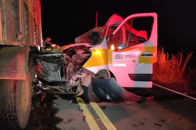 Veículo de emergência envolvido em um acidente com um caminhão, com danos visíveis na lateral do carro rodeado por equipes de resgate à noite.