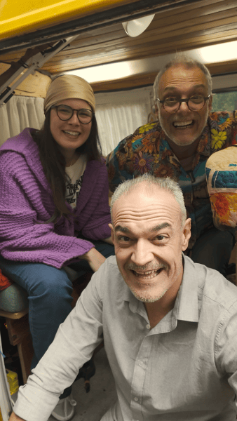 Três pessoas sorrindo dentro de uma van, uma mulher com suéter roxo e lenço na cabeça, um homem com óculos e camisa colorida, e outro homem com camisa clara.